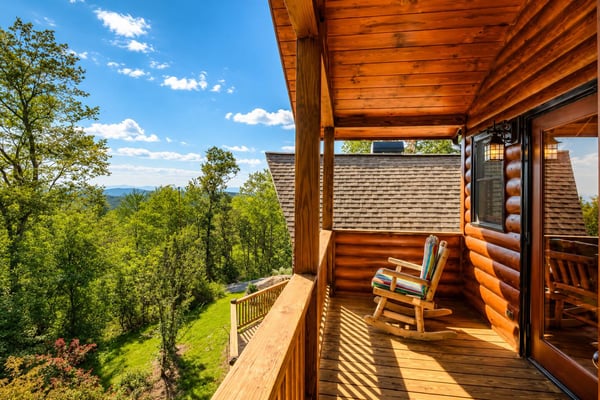 Log cabin deck with rocking chair overlooking forested mountains under blue sky